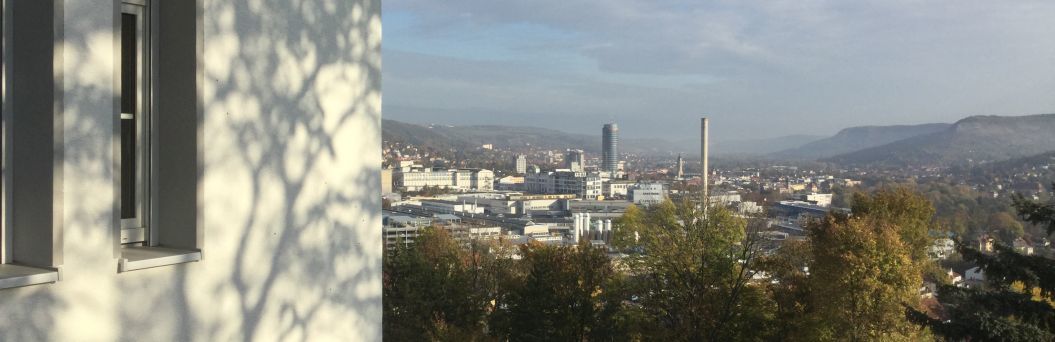 Zu sehen ist ein Teil des Gebäudes auf der linken Bildseite. Im Hintergrund sieht man die Stadt Jena.